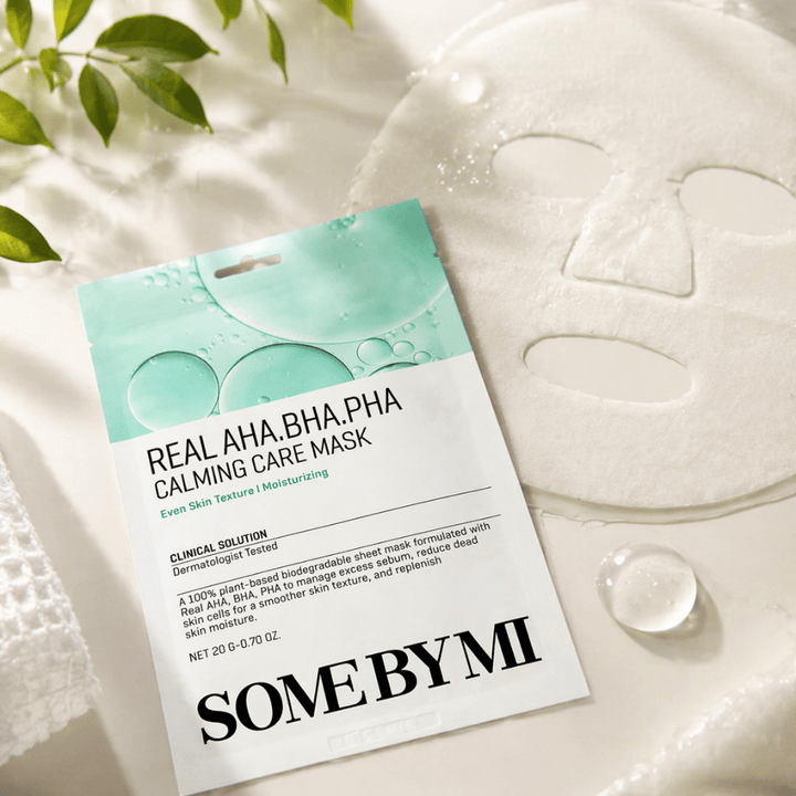 SOME BY MI Real AHA.BHA.PHA Calming Care Mask is shown together with the unfolded sheet mask. Around the product, moisture droplets and green leaves are visible, reinforcing the feeling of hydration, soothing care, and gentle exfoliation. The image highlights the mask's focus on a more even skin texture and improved moisture balance.