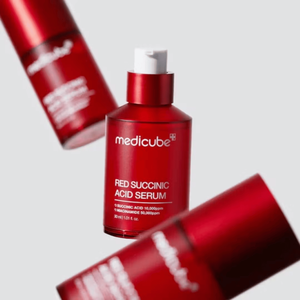 Medicube Red Succinic Acid Serum in red pump bottle, surrounded by more identical bottles against a light background. The product contains succinic acid and niacinamide to treat acne and pigmentation spots.