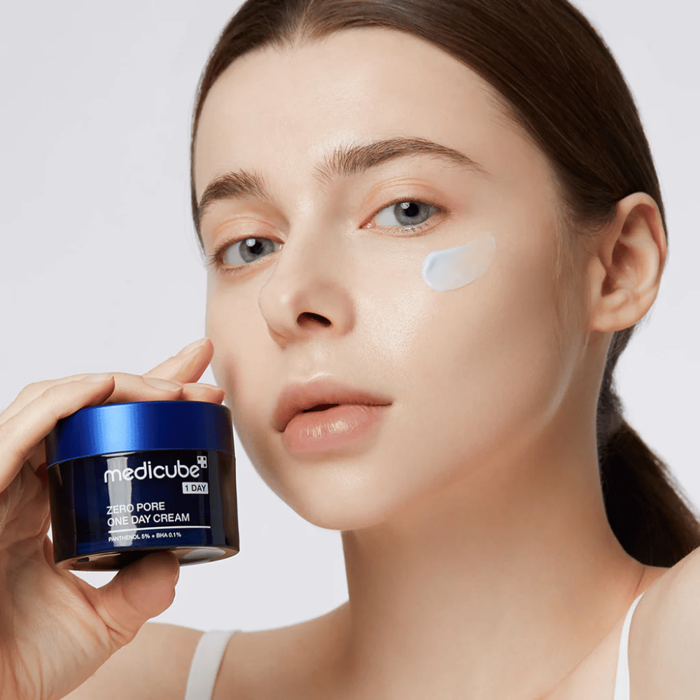 Woman with a natural look holds Medicube Zero Pore One Day Cream. A small amount of cream is applied to her cheek. The product is designed to improve skin texture and minimize pore size, with a light and moisturizing formula.