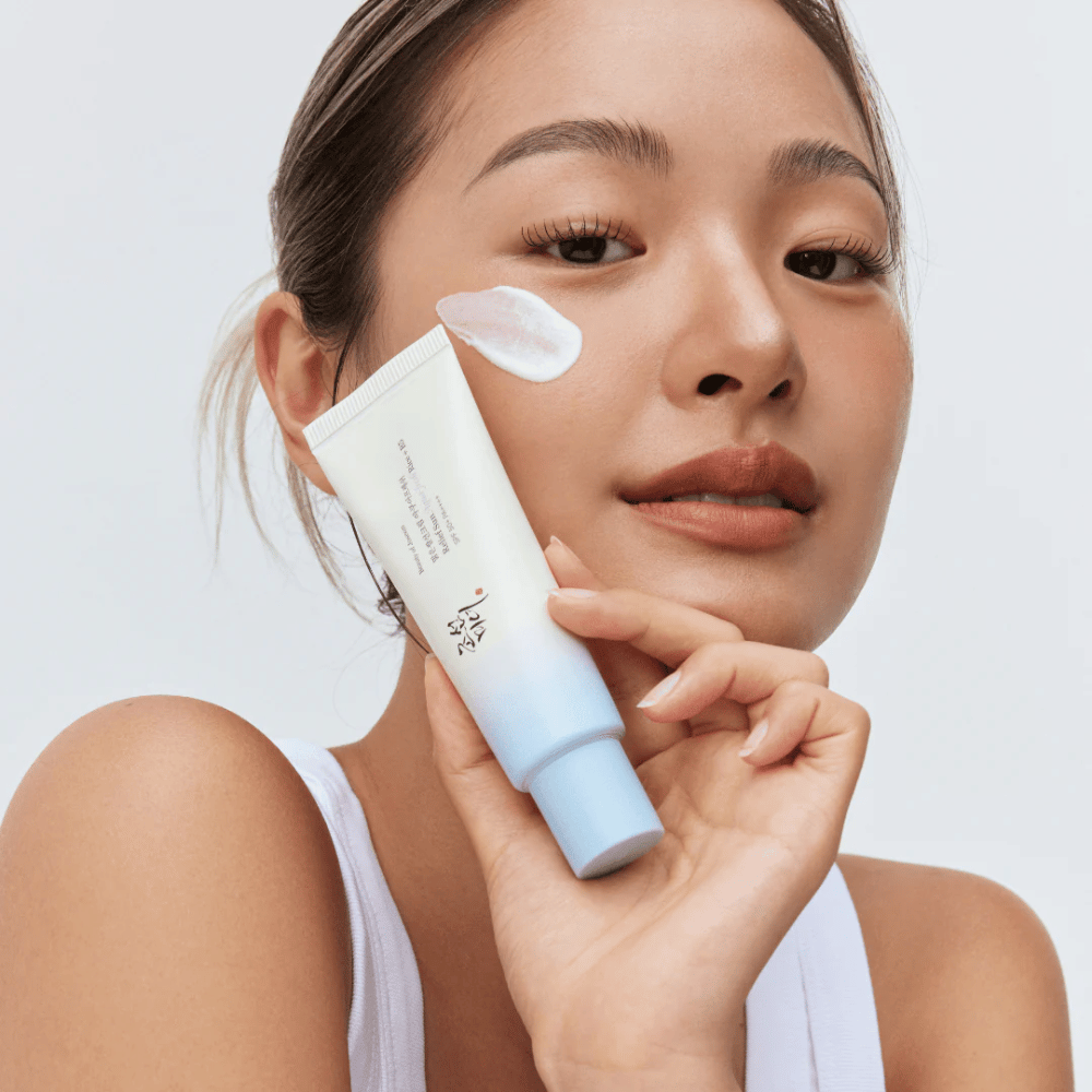 Woman holds Beauty of Joseon Relief Sun: Aqua-Fresh Rice + B5 sunscreen and shows its creamy texture on the cheek. A light sunscreen product that moisturizes and protects the skin with SPF 50+ PA++++.