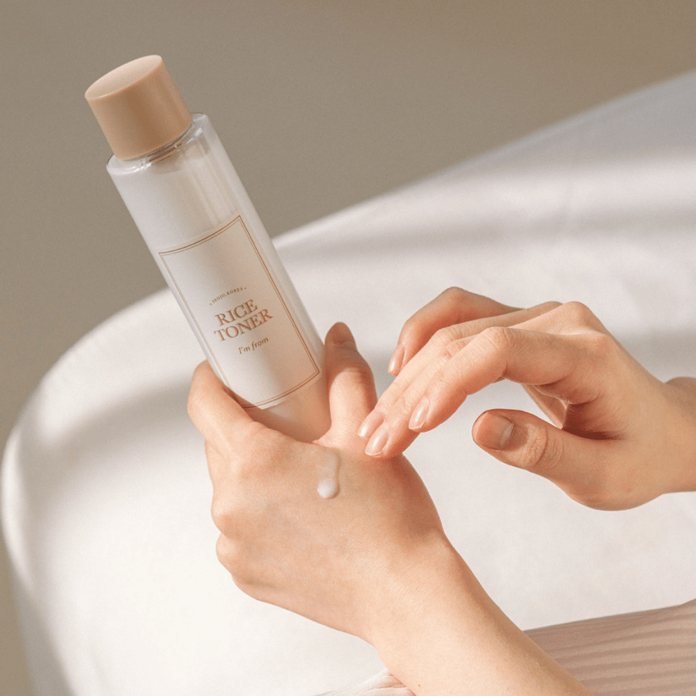 Hands holding a bottle of rice toner labeled "I'm from". A drop of the product is applied to one hand while the other hand prepares to apply it. The scene is softly lit with a warm tone.