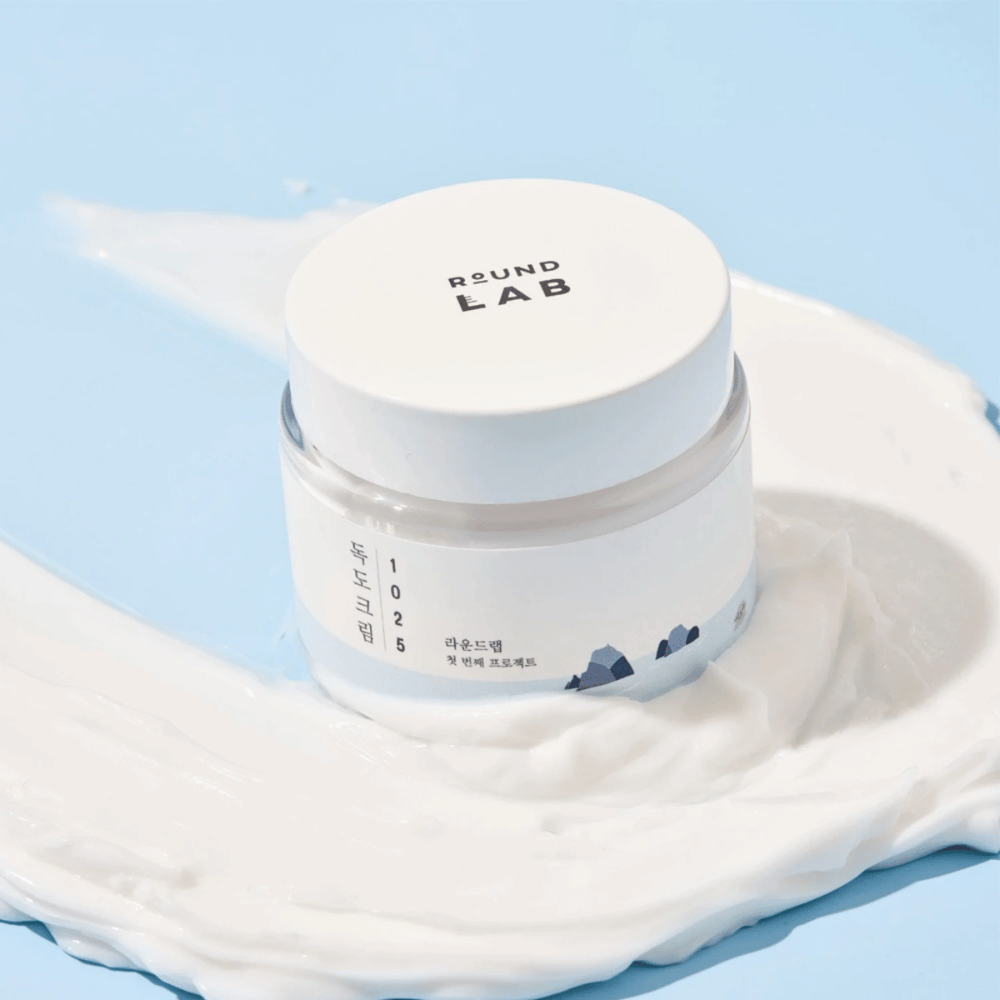 A jar with Round Labs 1025 Dokdo Cream 80 ml providing moisture, placed on a vibrant blue background.