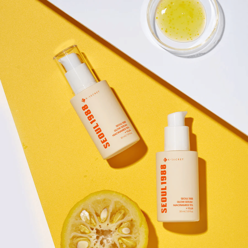 Two bottles of Seoul 1988 Glow Serum placed on a yellow background along with a bowl of yellow serum and a halved citrus fruit.