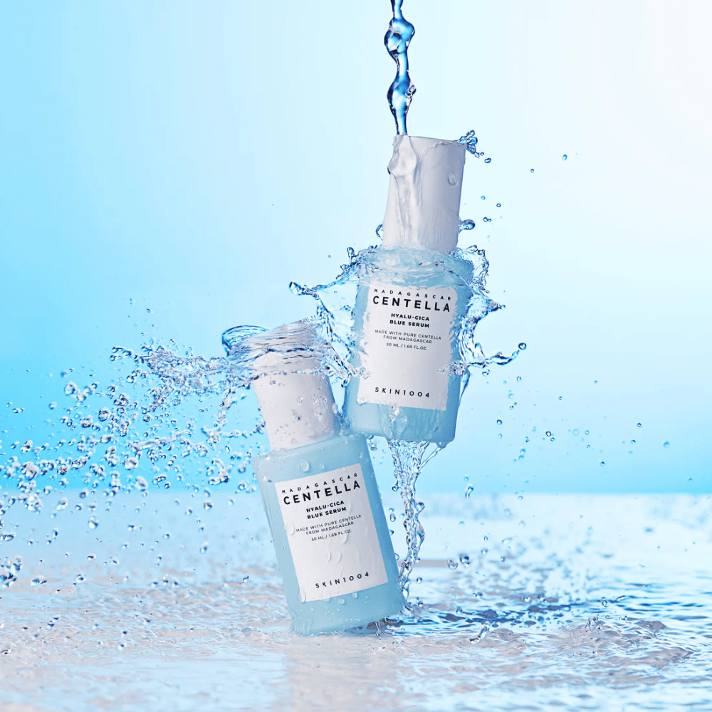Two bottles of SKIN1004 Madagascar Centella Hyalu-Cica Blue Serum are hit by a cascade of water against a light blue background.