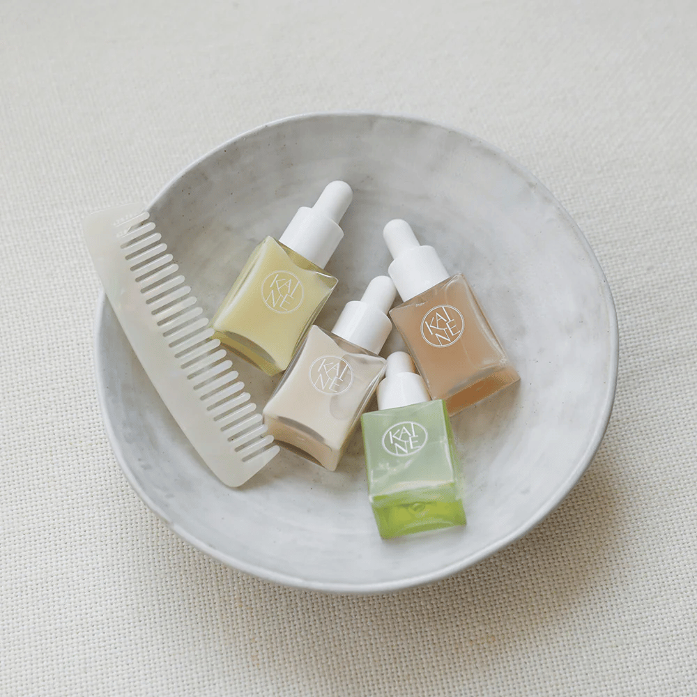 Four small serum bottles from KAINÉ Serum Discovery Kit lie in a white bowl next to a comb, with a natural and calm background in beige tone.