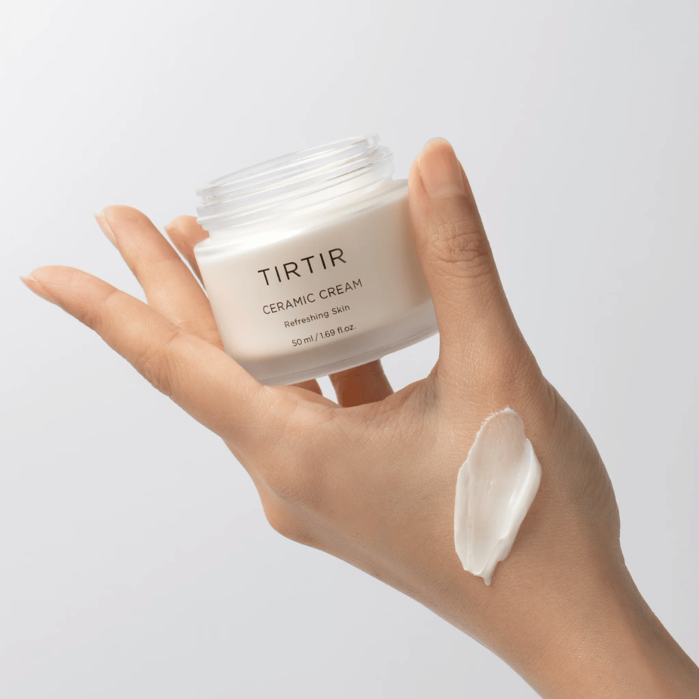 A hand holds a jar with TIRTIR Ceramic Cream. On the back of the hand, an amount of the cream has been applied. The cream appears to be thick and moisturizing. The packaging indicates that the content is for "Refreshing Skin" and the amount is 50 ml / 1.69 fl oz.