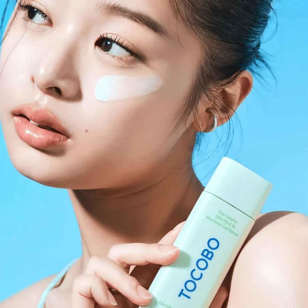 Model with sunscreen under the eye holding TOCOBO Cica Calming Sun Serum in mint green packaging against a blue background.