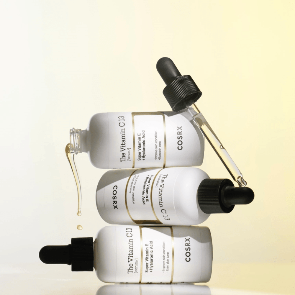 COSRX The Vitamin C 13 Serum with vitamin E and hyaluronic acid, three bottles with pipette and drops of Vitamin C serum for uneven skin tone and radiance.