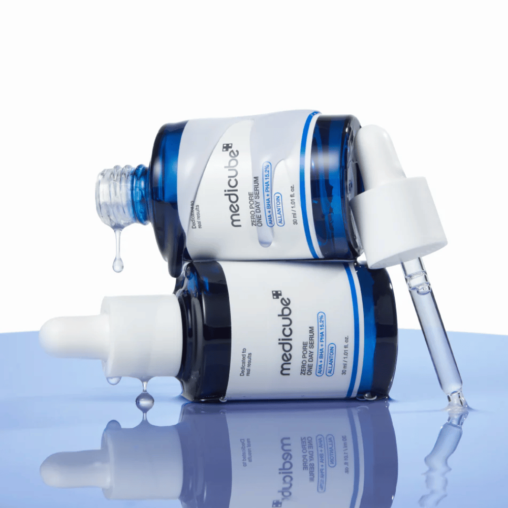 Two blue bottles of Medicube Zero Pore One Day Serum lying against each other, with pipettes and drops of serum on a reflective blue surface.