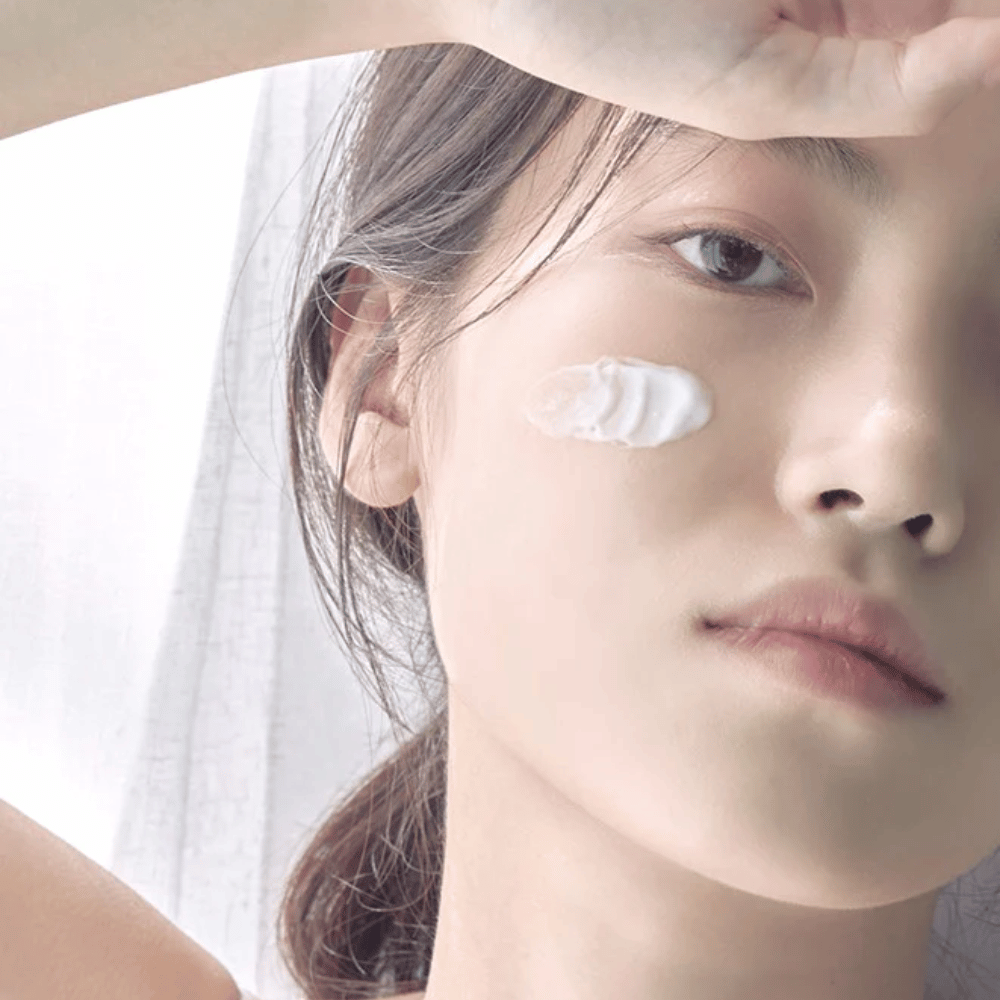 Close-up of a young woman with a dab of cream on her cheek and her hand raised above her forehead against a soft background.