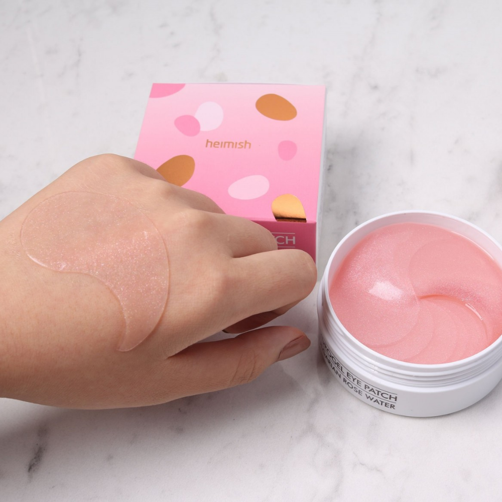 A hand holding a pink Bulgarian Rose Hydrogel Eye Patch 60pcs from heimish and applying eye mask.
